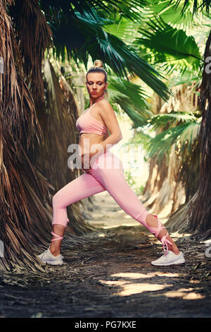 Young attractive blond woman in pink sportswear doing yoga pilates fitness exercises in tropical nature, holds hands on belly. Pre natal exercising, h Stock Photo