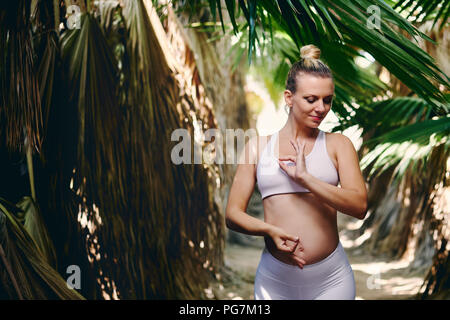 Young attractive blond woman in white sportswear doing yoga pilates fitness exercises in tropical nature. Pre natal exercising, healthy lifestyle, rec Stock Photo