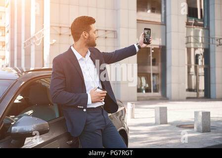 Visiting car dealership. Handsome man is doing selfie with his new car, showing thumb and smiling Stock Photo