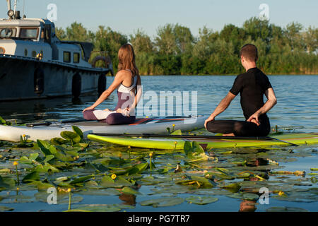 stand up paddle board yoga performed by beautiful couple on the bright background, yoga training on the beach Stock Photo