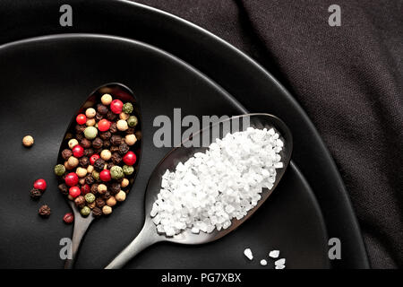 Close-up on salt and mixed pepper in dark spoons on black ceramic plates Stock Photo