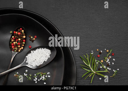 Salt in mixed pepper in dark spoons on black ceramic plates on slate stone with rosemary and thyme herbs Stock Photo