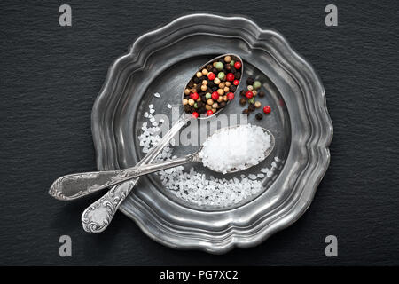 Salt in mixed pepper in vintage spoons on old metal plate on dark grey slate stone Stock Photo