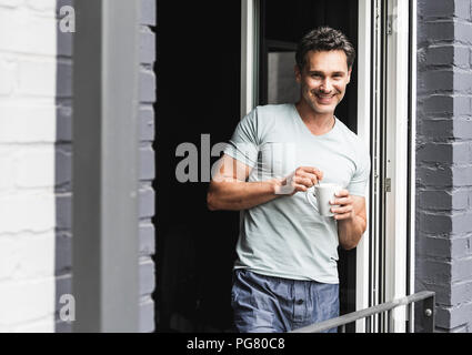 Portrait of smiling man in pyjama at home with cup of coffee at balcony door Stock Photo