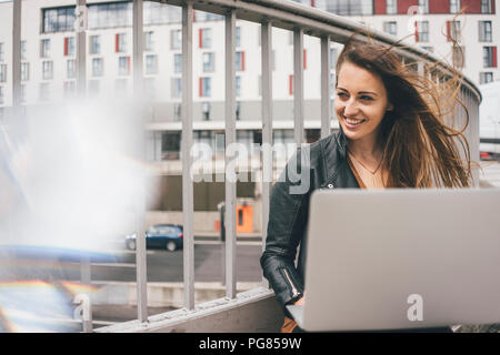 Happy young woman with windswept hair using laptop on motorway bridge Stock Photo
