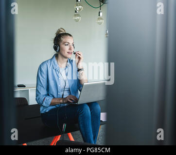 Young businesswoman sitting at desk, making a call, using headset and laptop Stock Photo