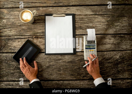 Hand of a Businessman on a Rustic Wooden Table with Tablet Computer, Calculator, Cup of Coffee and White Paper with Copy Space on a Clipboard. Stock Photo