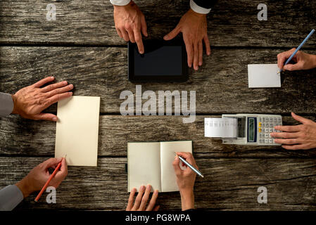 Top View of Businessmen Hands with Holding Notes, Tablet Computer and Printing Calculator on Top of a Rustic Wooden Table. Stock Photo
