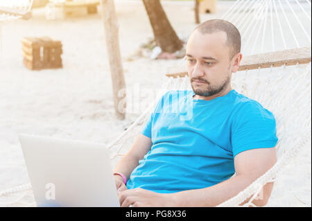 Man with a beard lying in a hammock with a laptop and looks at the screen. Work on the beach while traveling. The concept of remote work. Stock Photo