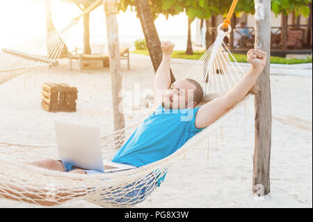 The man quite stretches lying in a hammock with a laptop. The concept of remote work. Stock Photo