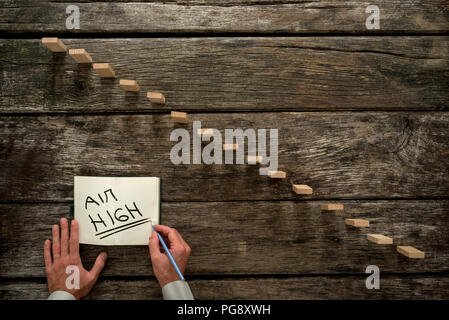 Top view of male hand writing an Aim high message on a white paper next to a steps made of wooden pegs resembling a staircase with textured oak backgr Stock Photo