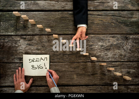 Top view of male hand writing an encouraging message Dream big in a notepad as a businessman walks his fingers up wooden pegs resembling staircase. Stock Photo