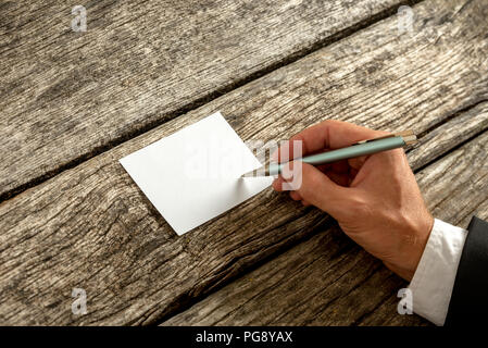 Overhead view of a hand in elegant suit writing on blank white card or paper with ballpoint pen on weathered old wooden desk, with copy space for you  Stock Photo
