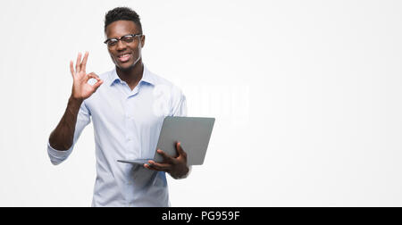 Young african american businessman using computer laptop doing ok sign with fingers, excellent symbol Stock Photo