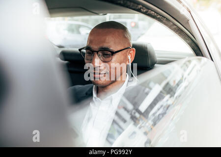 Business person wearing eyeglasses sitting in his sedan on back seat going to office. Man wearing earphones managing business work on the move sitting Stock Photo