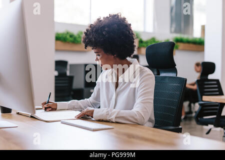Young african woman sitting at her desk and writing notes in her diary. Female executive working at her office desk. Stock Photo