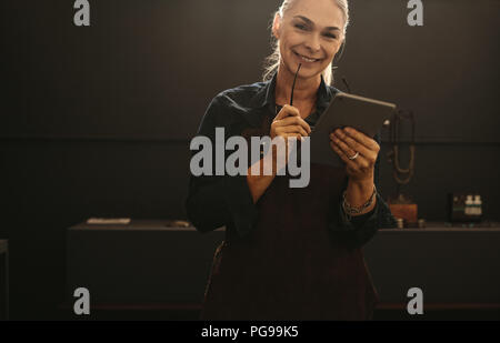 Portrait of smiling senior woman wearing apron holding digital tablet in her jewelry workshop. Smiling mature female goldsmith with tablet pc. Stock Photo