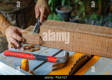 Man in apron chiseling wood with chisel on a table saw. Hammer laying on the table. Hands covered in saw dust. Carpentry tools, concept of wood work. Stock Photo
