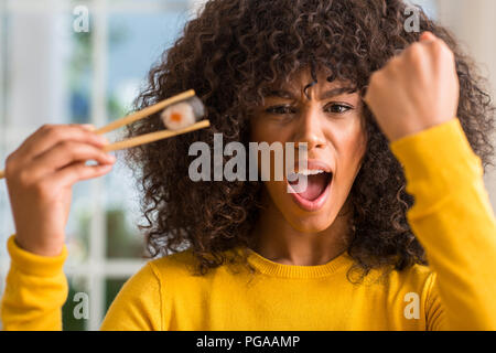 African american woman eating sushi using chopsticks at home annoyed and frustrated shouting with anger, crazy and yelling with raised hand, anger con Stock Photo