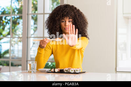 African american woman eating sushi using chopsticks at home with open hand doing stop sign with serious and confident expression, defense gesture Stock Photo