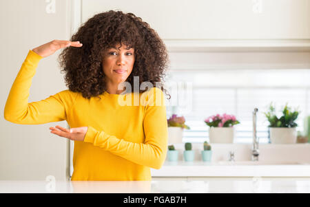 African american woman wearing yellow sweater at kitchen gesturing with hands showing big and large size sign, measure symbol. Smiling looking at the  Stock Photo