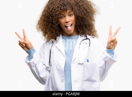African american doctor woman, medical professional working looking at camera showing tongue and making victory sign with fingers Stock Photo
