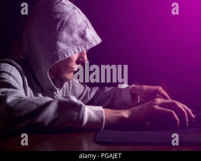 portrait of hooded man typing text on keyboard in front of pc monitor late in the night Stock Photo