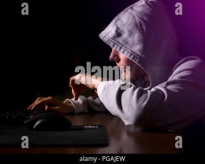 portrait of hooded man typing text on keyboard in front of pc monitor late in the night Stock Photo