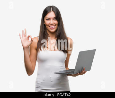 Young hispanic woman using computer laptop doing ok sign with fingers, excellent symbol Stock Photo