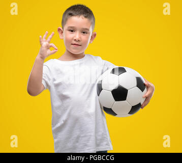 Dark haired little child playing with soccer ball doing ok sign with fingers, excellent symbol Stock Photo