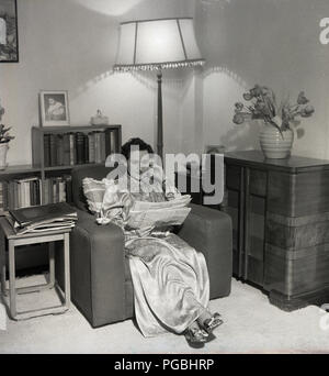 1950s, historical, picture shows a lady sitting in armchair in a lving room wearing a long dressing gown reading a newspaper, England, UK. Stock Photo