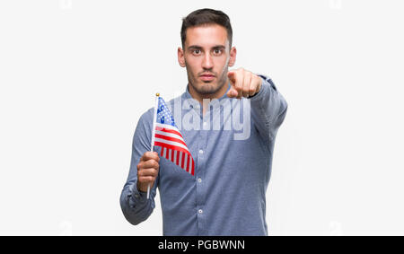 Handsome young man holding a flag of America pointing with finger to the camera and to you, hand sign, positive and confident gesture from the front Stock Photo