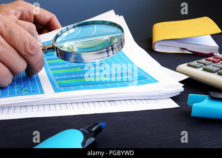 Man is holding magnifying glass above business documents with financial statistics. Audit concept. Stock Photo