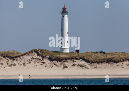 A couple walking on the beach as people stand on Lyngvig Lighthouse (Lyngvig Fyr) on the west coast of Jutland coast in Denmark. The lighthouse was built in 1906 and was the last built along this coast. Although no longer used for maritime navigation, it was built following a tragic accident at sea in 1903, where 24 sailors lost their lives. The light was lit for the first time on 3rd November 1906. Stock Photo