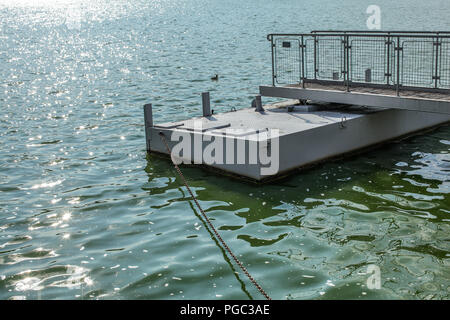 Metal boat pier, on a green water lake, sun sparkling on small waves, chain leading to shore. Brno, Czechia Stock Photo