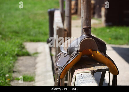 Old worn horse saddle laying on wooden fence lit by sun. Stock Photo