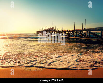 Sea sunrise near a old pier. Calm waves and soft foam on the beach sand spark in the sun rays. Stock Photo