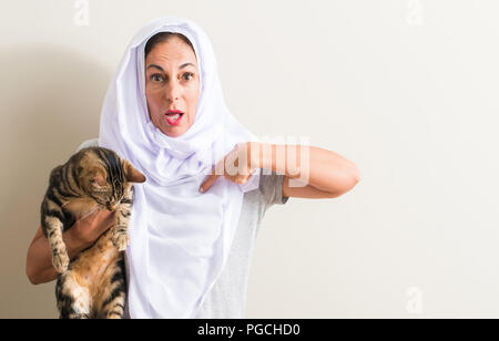 Arabian woman wearing white hijab holding a cat with surprise face pointing finger to himself Stock Photo