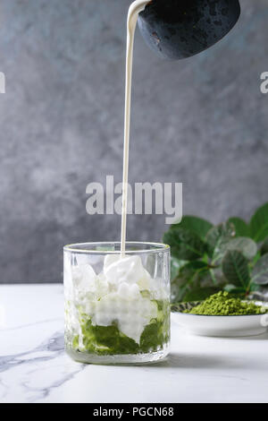 Cream pouring from jug to matcha green tea iced latte or cocktail in glass, with ice cubes, matcha powder on white marble table, decorated by green br