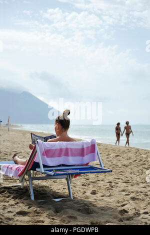 Woman at the beach relaxes by reading a gossip magazine. On the background a couple of lovers is walking. Sabaudia, Lazio, Italy Stock Photo