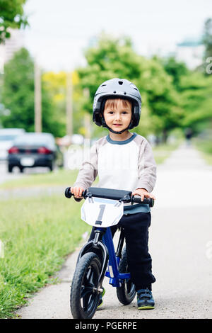 Portrait of smiling little boy toddler riding a balance bike bicycle in helmet on the road outside outdoors on spring summer day, seasonal child activ Stock Photo