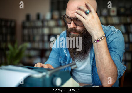 Thoughtful bearded stylish writer typing on typewriter. Modern writer in glasses working on new book in library Stock Photo