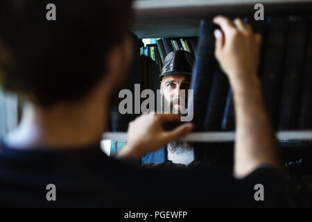 Man taking book from library bookshelf and sees bearded man. Peeping man in black hat among bookshelves Stock Photo