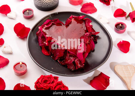 Romantic heart-shaped red cake with rose petals served on a white table decorated with candles,, rose petals and hearts for an anniversary or Valentin Stock Photo