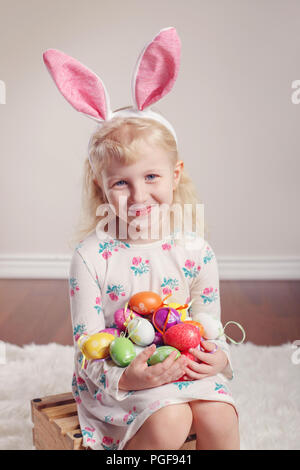Cute adorable Caucasian child girl wearing Easter bunny rabbit ears sitting on wooden box in studio indoors. Kid holding holiday colorful eggs celebra Stock Photo