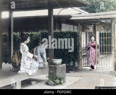 [ c. 1890s Japan - Washing Hands ] —   A woman in kimono uses a Hishaku (ladle) to splash water over the hands of another woman who is washing her hands. The two women are sitting on the porch of a private house. A third woman is entering the garden through the gate. Washing hands (Temizu) is an important purification ritual when entering shinto shrine grounds, but was also done upon entering a private home.  19th century vintage albumen photograph. Stock Photo