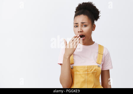 Hate boring people. Indifferent tired and bored arrogant african-american female with curly hair in yellow overalls, yawning and covering opened mouth with palm, being careless and ininterested Stock Photo