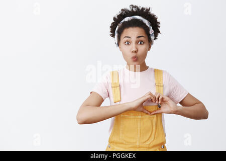 Human needs love. Portrait of hopeful cute and tender African American in yellow overalls and headband, showing heart gesture near chest and folding lips in kiss, posing over gray background Stock Photo