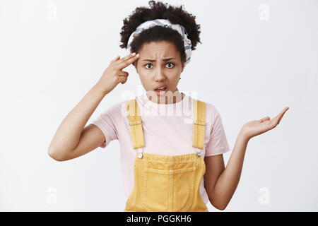 You lost your mind. Portrait of confused and displeased disappointed african american woman in headband and yellow overalls, holding finger near temple, shrugging, looking at crazy or weird friend Stock Photo