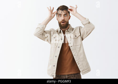 Portrait of stunned speechless and shocked good-looking mature bearded guy taking off glasses to react on something stunning and shook popping eyes and opening mouth from stupor over gray wall Stock Photo